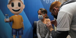 Ein Mädchen wird geimpft, eine kleine Figur mit Impfflasche ist auf eine blaue Wand gemalt