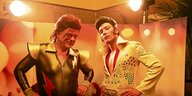Zwei Männer stehen vor einer Kulisse. Einer hat sich als David Bowie verkleidet, der andere als Elvis.
