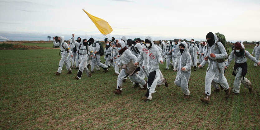 In weißen Anzügen setzen sich Demonstranten in Bewegung, einer schwingt eine gelbe Fahne auf einem Feld