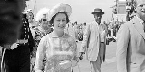 Eine Frau mit Hut, Brosche und Handtasche umringt von uniformierten Würdenträgern