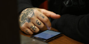 Eine Hand an einem Smartphone, auf den Fingern sind die Buchstaben frei taetowiert