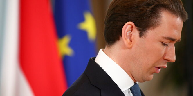 Kurz im Profil, er guckt herab. Hinter ihm sind Teile der österreichischen Flagge sowie die Fahne der EU zu sehen