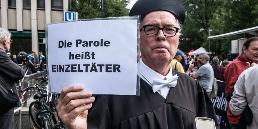 Mensch in Anwaltskluft und einem Schild mit der Aufschrift "Die Parole heißt Einzeltäter"