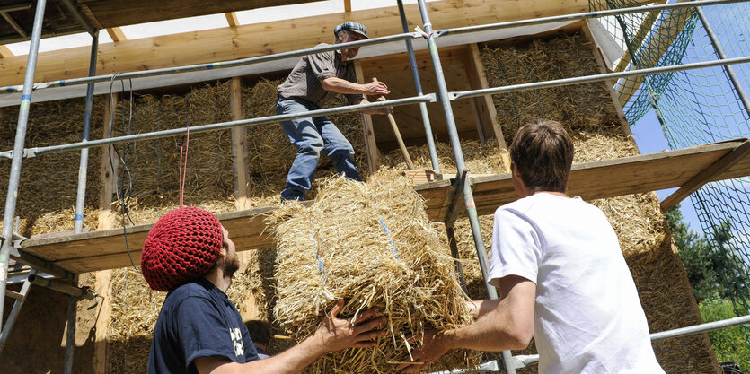 Zwei Leute reichen einem dritten einen Strohballen hinauf, der in der Fassade eines neues Hauses verbaut werden soll