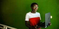 Tharcissie Idufashesteht mit aufgeklapptem Laptop vor einer grünen Wand
