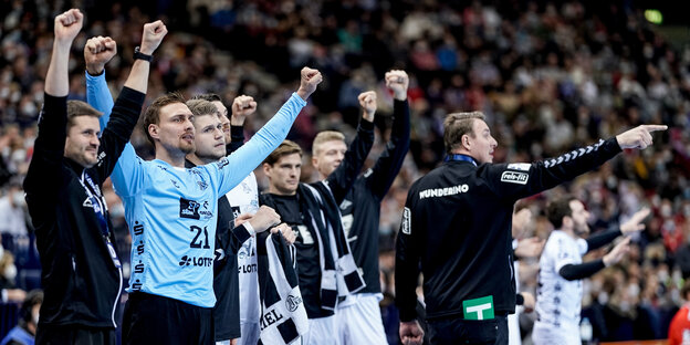 Auswechselspieler des THW Kiel stehen mit nach oben gehaltenen Armen am Spielfeldrand. Der Trainer zeigt in die Richtung rechts aus dem Bild