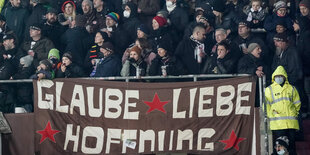 Fußballfans stehen dicht aneinander, im Vordergrund ein Transparent mit den Worten "Glaube, Liebe, Hoffnung"