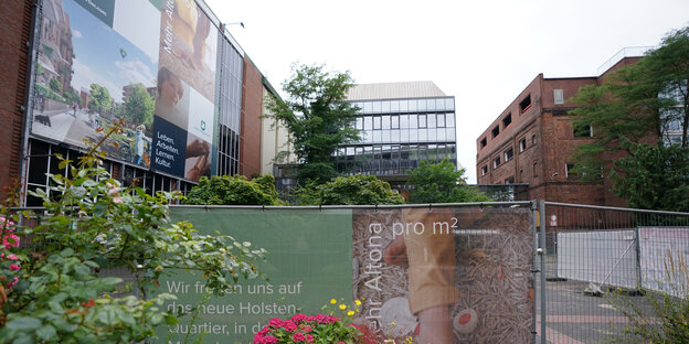 Blick über einen Zaun auf eine Gebäudeflucht, links mit großen Plakaten versehen