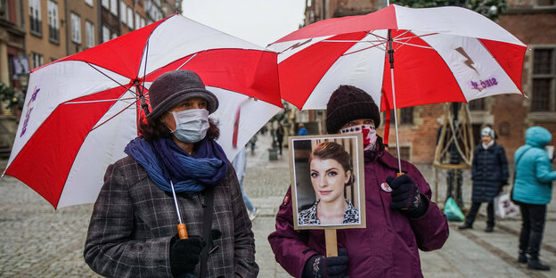 Frauen mit Mundschutz uter regenschirmen in den polnischen Nationalfarben rot und weiß. Eine hält ein Foto in der Hand