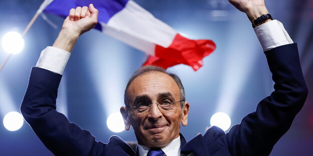 Eric Zemmour hebt die Hände, im Hintergrund schwenkt jemand die Frankreich-Fahne