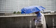 Ein Mann trägt eine blauen Schirm mit dem Logo der EU und geht am Gebäude der Europäischen KOmmission entlang