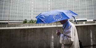 Ein Mann trägt eine blauen Schirm mit dem Logo der EU und geht am Gebäude der Europäischen KOmmission entlang