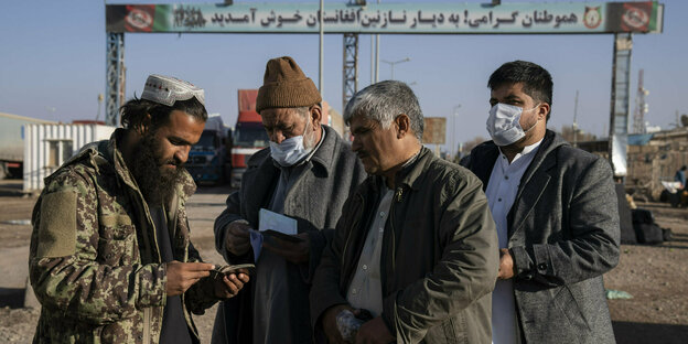 Ein Mann kontrolliert Dokumente dreier Männer, im Hintergrund ein Grenzübergang