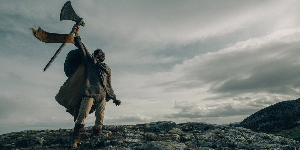 Gawain (Dev Patel) hebt im Gebirge seine Axt in die Höhe.