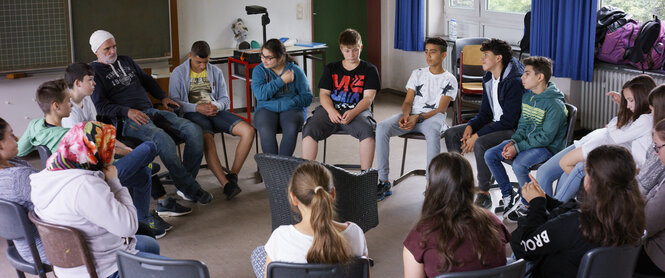 Herr Bachmann sitzt mit seinen Schülern im Klassenzimmer im Kreis.