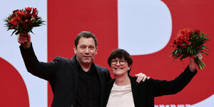 Lars Klingbeil und Saskia Esken halten Blumensträuße in die Höhe, vor einer Wand mit einem riesigen SPD-Logo