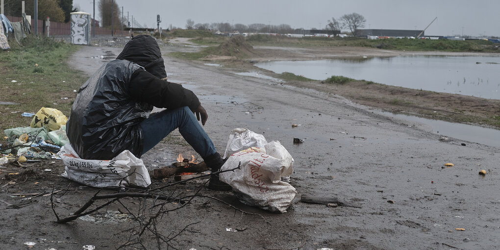 Eine Person ist in einen Plastiksack gehüllt und sitzt auf dem nassen Boden und macht ein Feuer