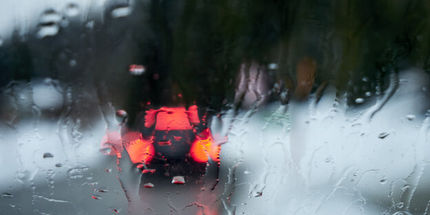 Hinter einer Autoscheibe ist durch den Regen verschwommen eine Straße zu sehen auf der ein Auto fährt. Daneben liegt Schnee.