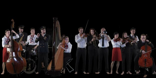 12 Musiker:innen stehen mit Bass, einer Harfe und anderen Instrumenten nebeneinander vor einem schwarzen Hintergrund