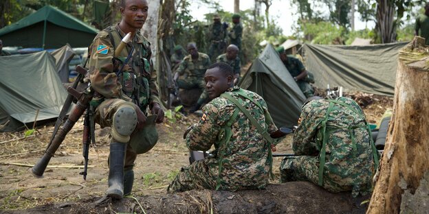 Soldaten sitzen auf einem Baumstumpf, einer steht und stützt sein Bein auf den Holzstumpf, dahinter Zelte