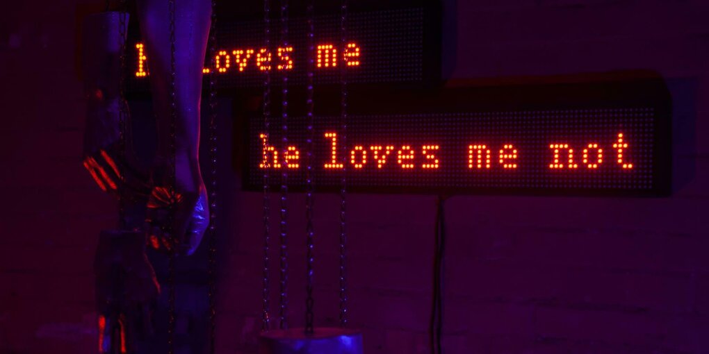 Auf einem rot erleuchteten Neonschriftband erscheinen die Sätze "He loves me" und "He loves me not"