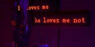 Auf einem rot erleuchteten Neonschriftband erscheinen die Sätze "He loves me" und "He loves me not"