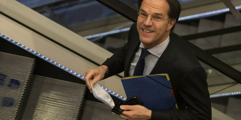 Mark Rutte schaut auf einer Rolltreppe nach oben und hat eine Maske in der Hand