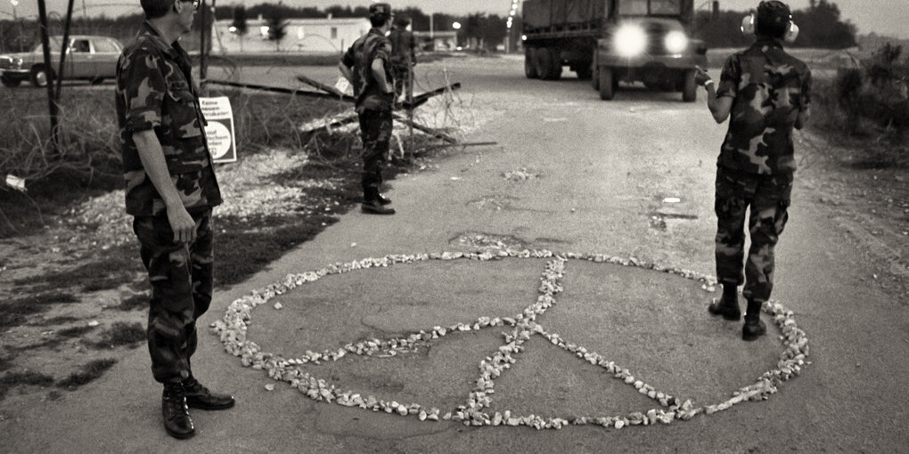 Ein aus Steinchen gelegtes Peace-Zeichen am Boden zwischen Männern in Uniform, ein LKW fährt auf sie zu