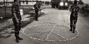 Ein aus Steinchen gelegtes Peace-Zeichen am Boden zwischen Männern in Uniform, ein LKW fährt auf sie zu