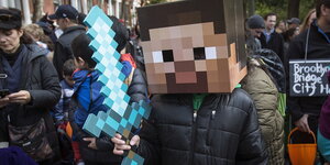Ein Kind ist als ein Charakter des Videospiels Minecraft an Halloween verkleidet. Es hat eine große, breite Maske auf, die ein verpixeltes Gesicht zeigt. In der Hand hält es ein Schwert aus Pappe. Im Hintergrund sind viele Menschen zu sehen