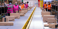 Mitarbeiter mit orangen und pinkfarbenen Westen sortieren am Fließband Amazon-Pakete