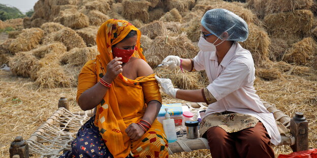 Eine Frau mi torangem Sari wird vor einem Stapel Heuballen geimpft