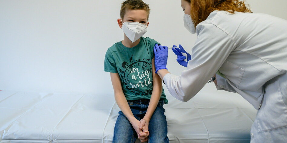 Ein Junge wird geimpft, er faltet die Hände und sitzt auf einer Liege, er trägt ein grünes T-Shirt mit der Aufschrift "it`s a big world"