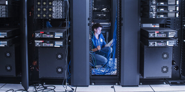 Eine Frau hockt in einem Serverraum zwischen Kabeln und Rechnern