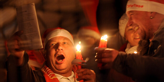 Ein Herr mit Fußballschal sing lauthals ein Weihnachtslied, ein Mann daneben hält eine Kerze und lacht