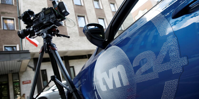 Ein Auto mit der Aufschrft TVN24 steht vor einem Haus. Neben dem Fahrzeug steht eine Filmkamera auf einem Stativ