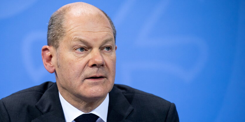 Olaf Scholz, ein alter Mann mit einer Glatze in der MItte des Kopfes, blickt an der Kamera vorbei. Er trägt einen Anzug. Hinter Scholz eine blaue Wand, auf der eine Kralle des Bundesadlers zu erkennen ist