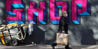 Ein Mann mit einer Primark-Tüte geht an einem Graffito mit dem Schriftzug "Shop" vorbei.