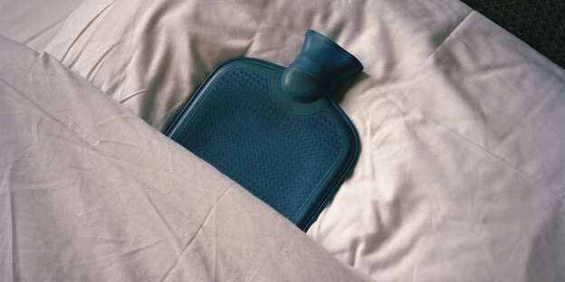 blaue Wärmeflasche liegt auf dem Kopfkissen eines weiß bezogenen Betts
