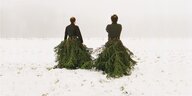 Zwei Männer gehen durch den Schnee, sie tragen schwarze Pullover und haben lange Röcke aus Tannebäumen, die sie über die Erde ziehen