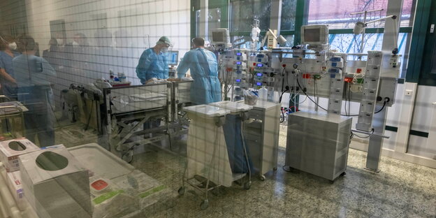 Zwei Personen in blauen Kitteln stehen hinter einer Glasscheibe in einem Behandlungszimmer einer Intensivstation am Bett einer kranken Person. Überall sind technische Geräte. Im Fenster spielgen sich weitere Personen.
