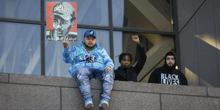 Drei Junge Männer an einer niedrigen Mauer. Einer der Männer sitzt auf der Mauer und hält ein Plakat mit dem Gesicht von Daunte Wright in die Luft. Die anderen beiden stehen hinter der Mauer. Einer streckt die rechte Faust in die Luft.