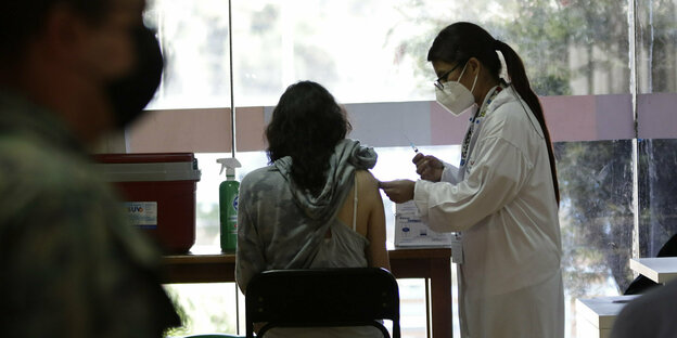 Eine Frau sitzt auf einem Sthul mit dem Rücken zur Kamera. Eine Ärztin in Kittel und mit Mundschutz steht neben ihr und gibt ihr eine Impfung in den rechten Oberarm.