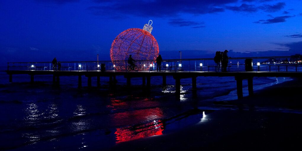 Eine Lichtinstallation in Form einer großen roten Christbaumkugel leuchtet hinter einem Steg auf der Wasseroberfläche