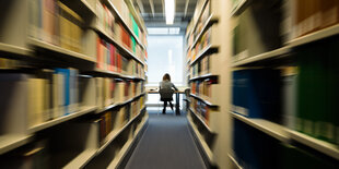 Zwei volle Bücherregale bilden einen Gang. An dessen Ende sitzt eine Studentin am Schreibtisch.