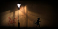 Eine Straßenlaterne beleuchtet eine dunkle Straße, rechts von ihr läuft eine Frau im Dunkeln daran vorbei.