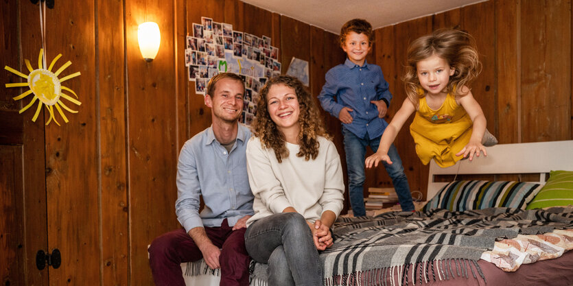 Junges Paar sitzt auf einem Bett, auf dem zwei kleine Kinder hüpfen