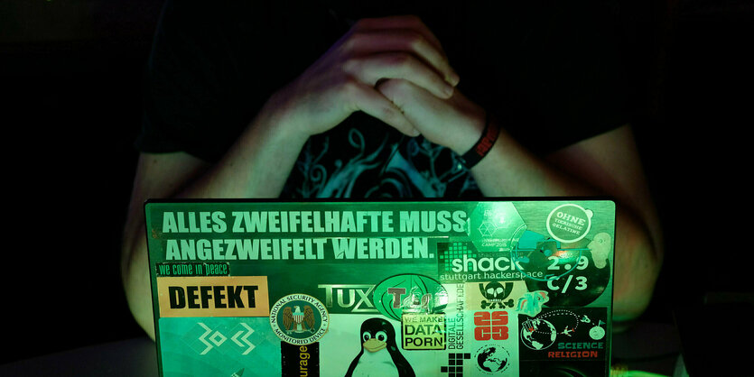 Eine Person hinter einem Laptop, auf dem verschiedene Sticker geklebt sind. Unter anderem steht auf einem Sticker "Defekt", ein anderer zeigt das Linux-Maskottchen Tux, ganz oben steht auf einem Sticker: "Alles Zweifelhafte muss angezweifelt werden."