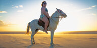Eine mollige Frau sitzt selbstbewußt auf einem Pferd im Badedress an einem Strand im warmen Sonnenlicht