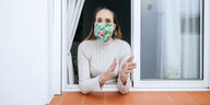eine Frau mit bunter Gesichtsmaske steht am offenen Fenster und klatscht